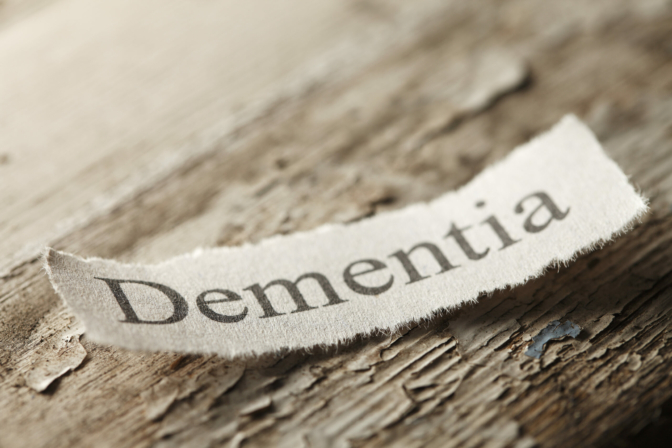 The word 'Dementia" on a torn piece of paper that rests on an old weathered piece of wood.  The old paint is flaking off of the wood surface to help convey the feeling of age.